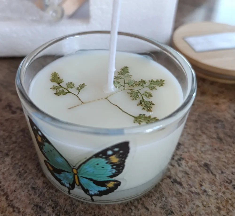 Bougie artisanale blanche dans un verre transparent de 110 grammes, décoré d’un papillon bleu, avec une petite fougère séchée incrustée dans la cire près de la surface.