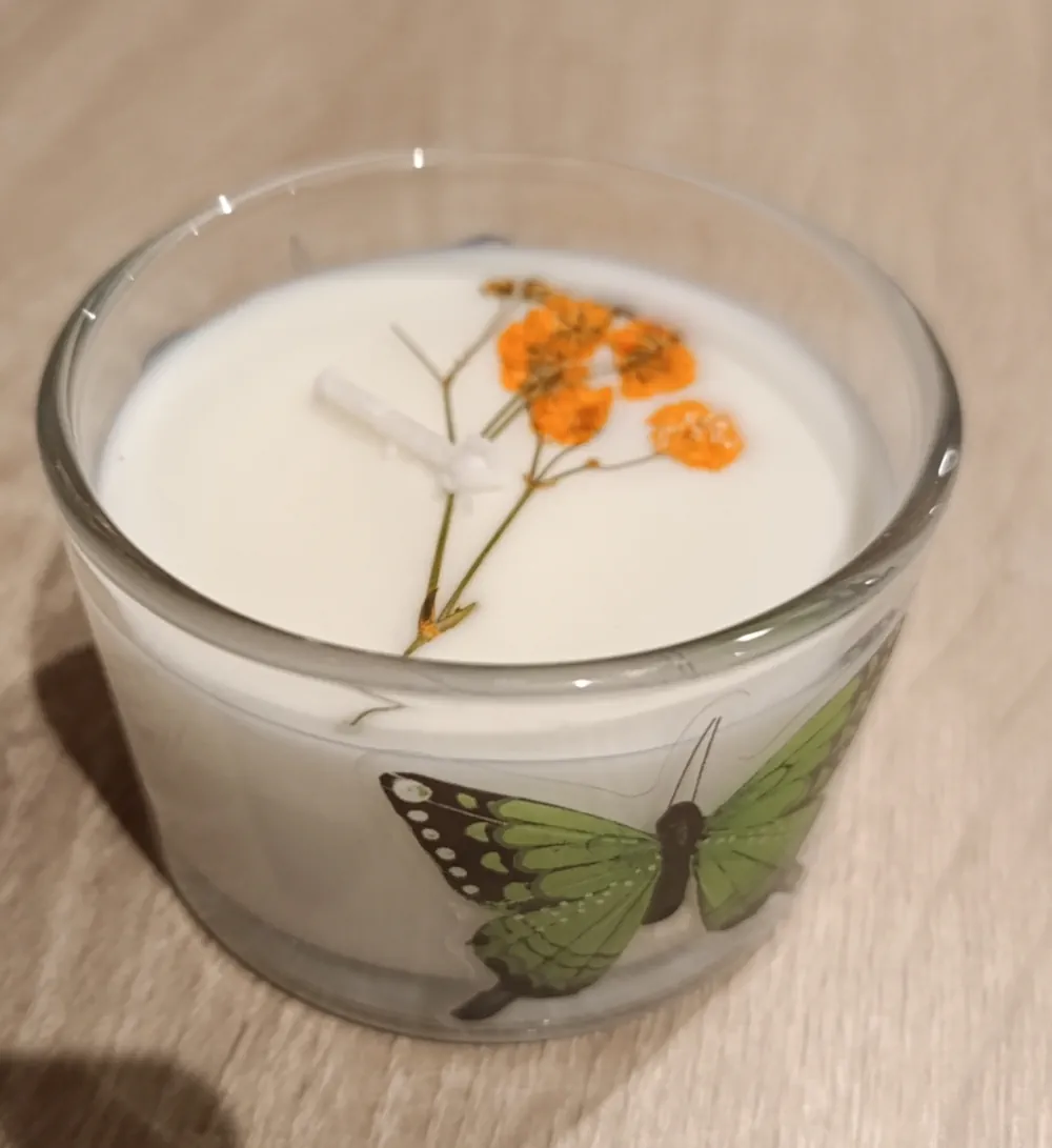 Bougie blanche artisanale dans un verre transparent de 110 grammes, décoré d’un papillon vert, ornée de petites fleurs séchées orange incrustées dans la cire autour de la mèche.
