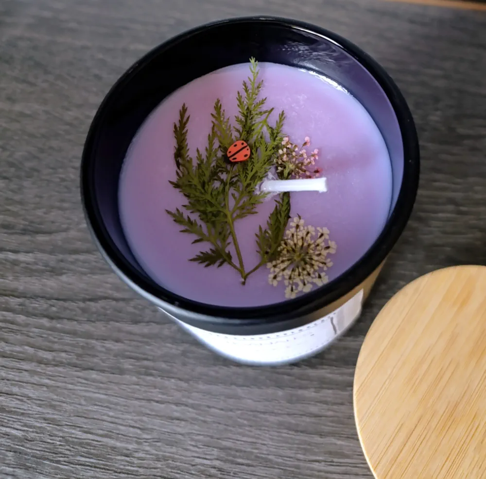 Bougie artisanale à la cire mauve décorée de fleurs séchées, d'un feuillage de fougère et d'une petite figurine de coccinelle dans un contenant noir de 200ml, parfum Lilas.