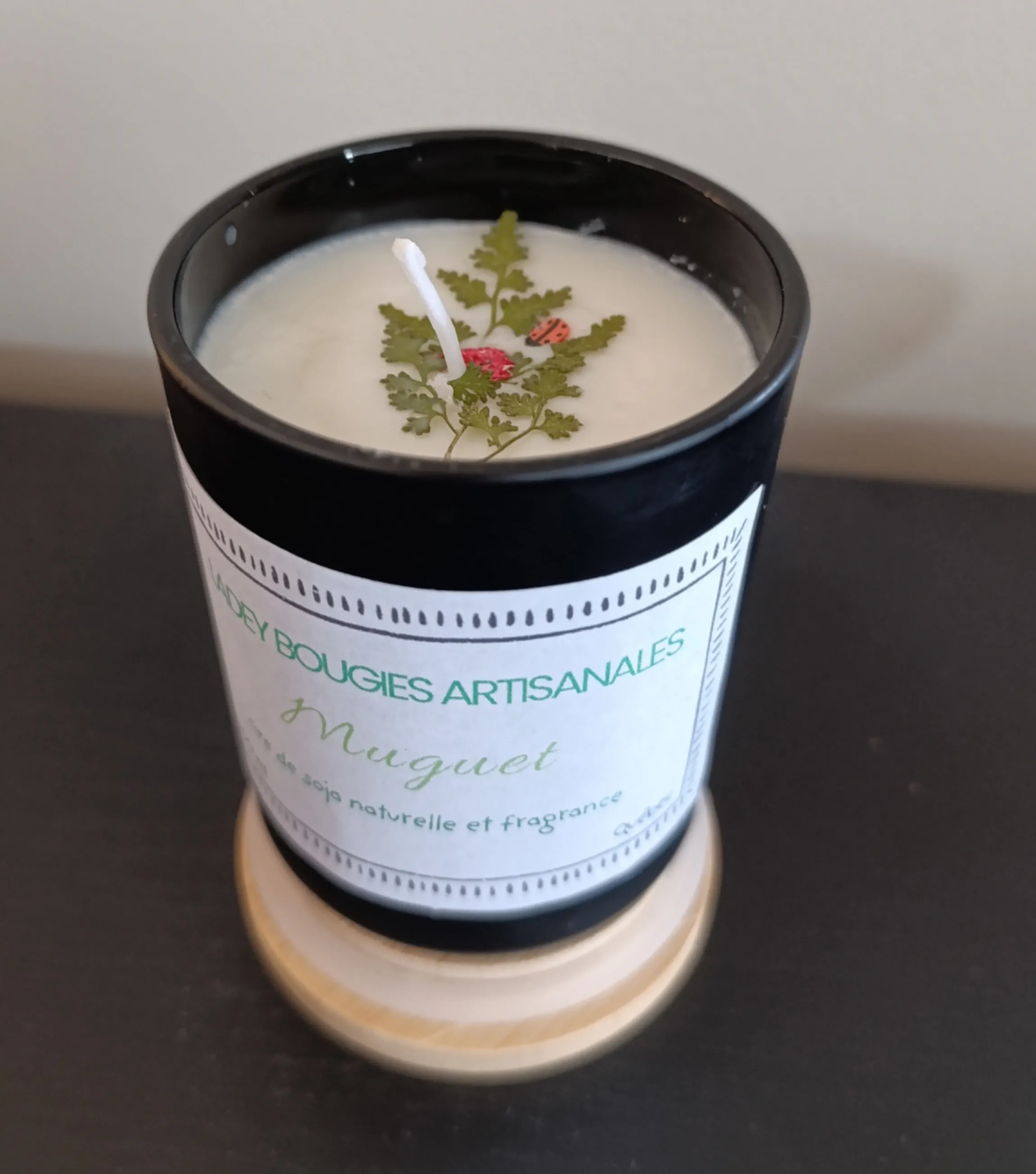 Bougie artisanale dans un contenant noir de 200ml, décorée d’une feuille verte et de petits ornements rouges, parfum Muguet.