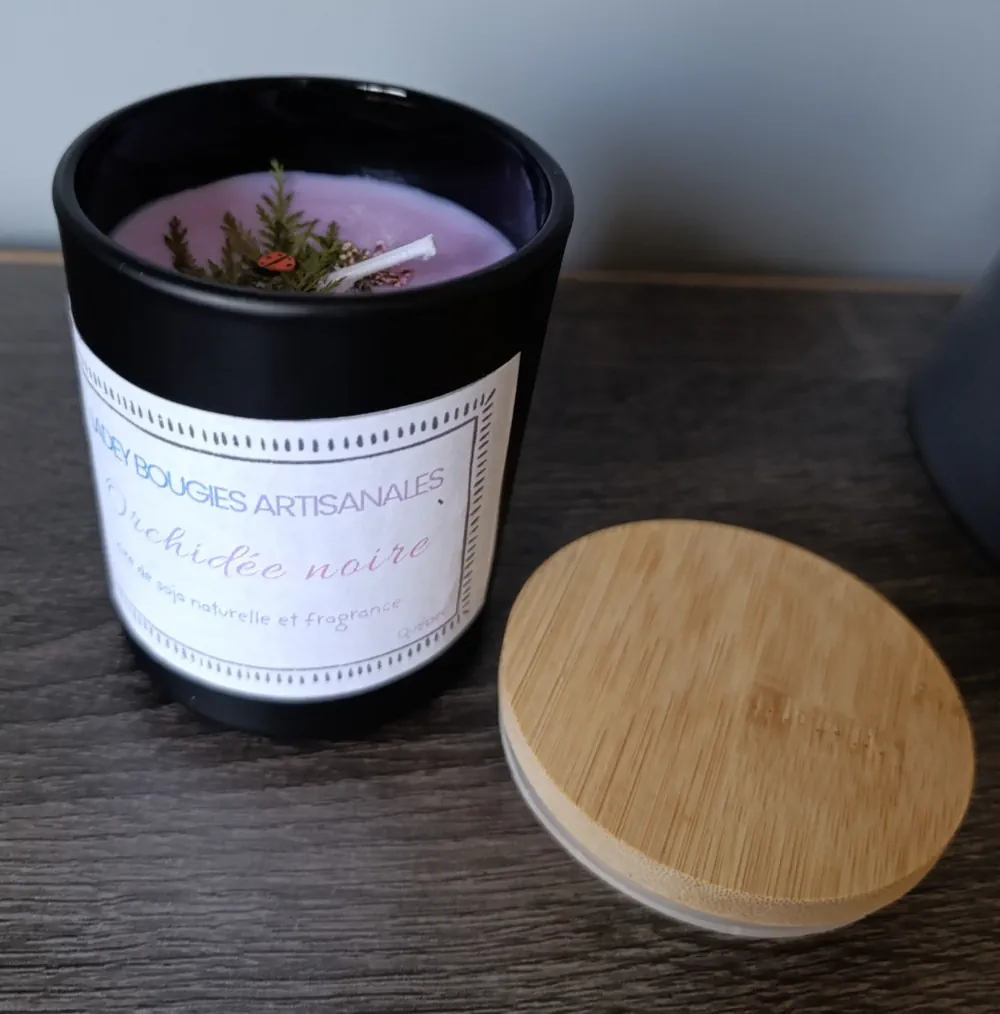 Bougie artisanale dans un contenant noir de 200ml, à la cire mauve décorée de feuillage vert et d’un petit ornement rouge, parfum Orchidée noire.