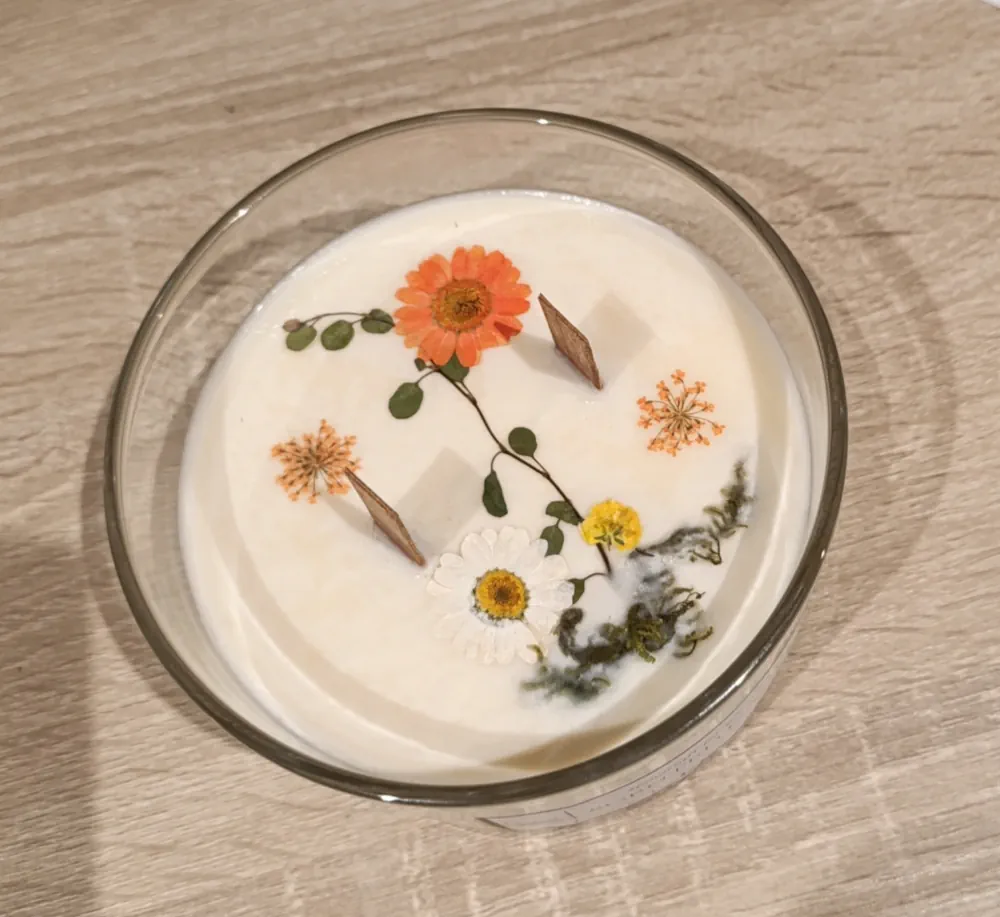 Bougie artisanale en cire blanche et mèches en bois dans un récipient en verre transparent de 110 grammes, décorée de fleurs séchées oranges, de marguerites séchées et de fleurs jaune et feuilles vertes séchées.