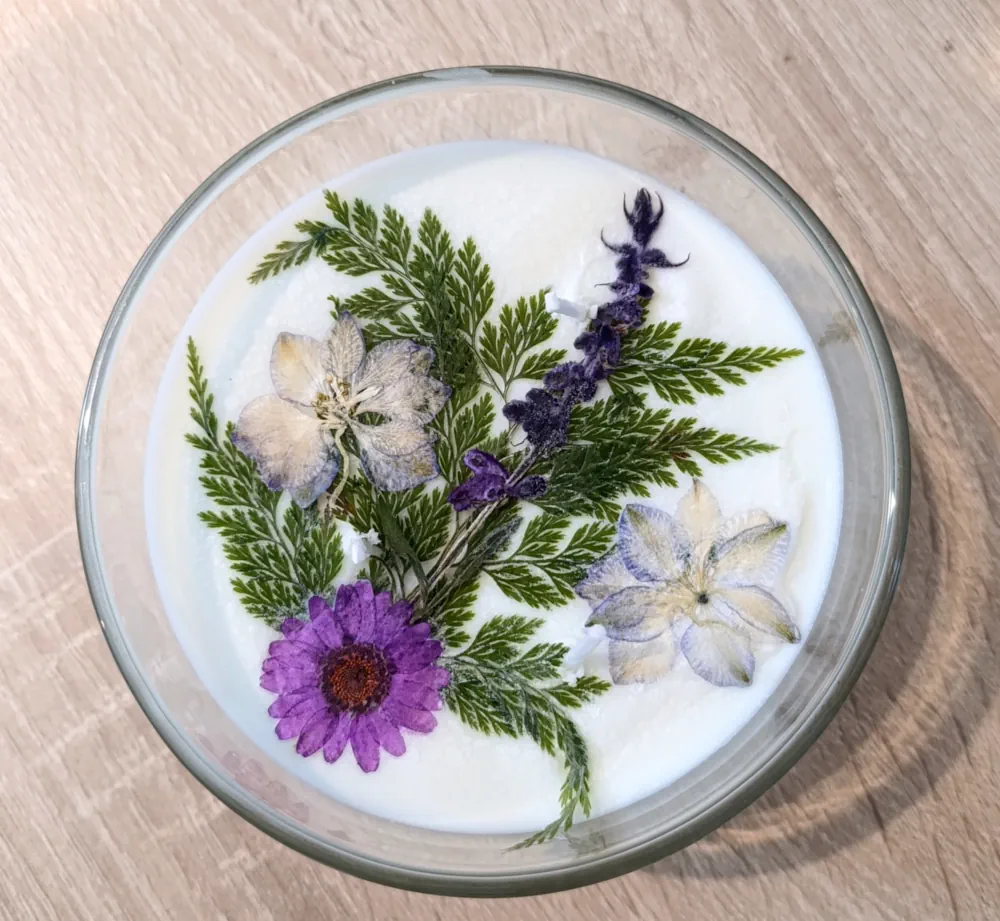 Bougie artisanale blanche dans un récipient en verre transparent de 110 grammes, décorée en surface de petites fleurs séchées violettes et lavande, de fleurs crème délicates et de frondes de fougère vertes soigneusement disposées.