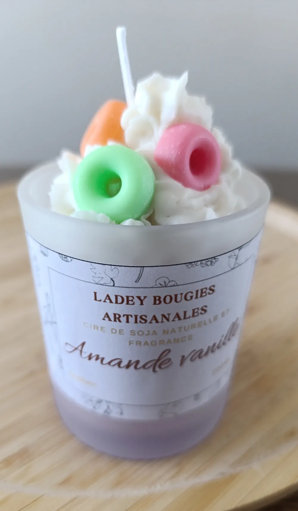 Bougie artisanale dans un verre givré de 200ml au parfum amande et vanille, surmontée d’un tourbillon de cire blanche façon crème fouettée et décorée de trois minis beignes (donuts) colorés en cire — vert, rose et orange.