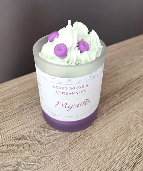 Bougie artisanale dans un verre givré de 200ml au parfum myrtille, surmontée d’un tourbillon de cire blanche façon crème fouettée et décorée de petites billes de cire violette évoquant des myrtilles.