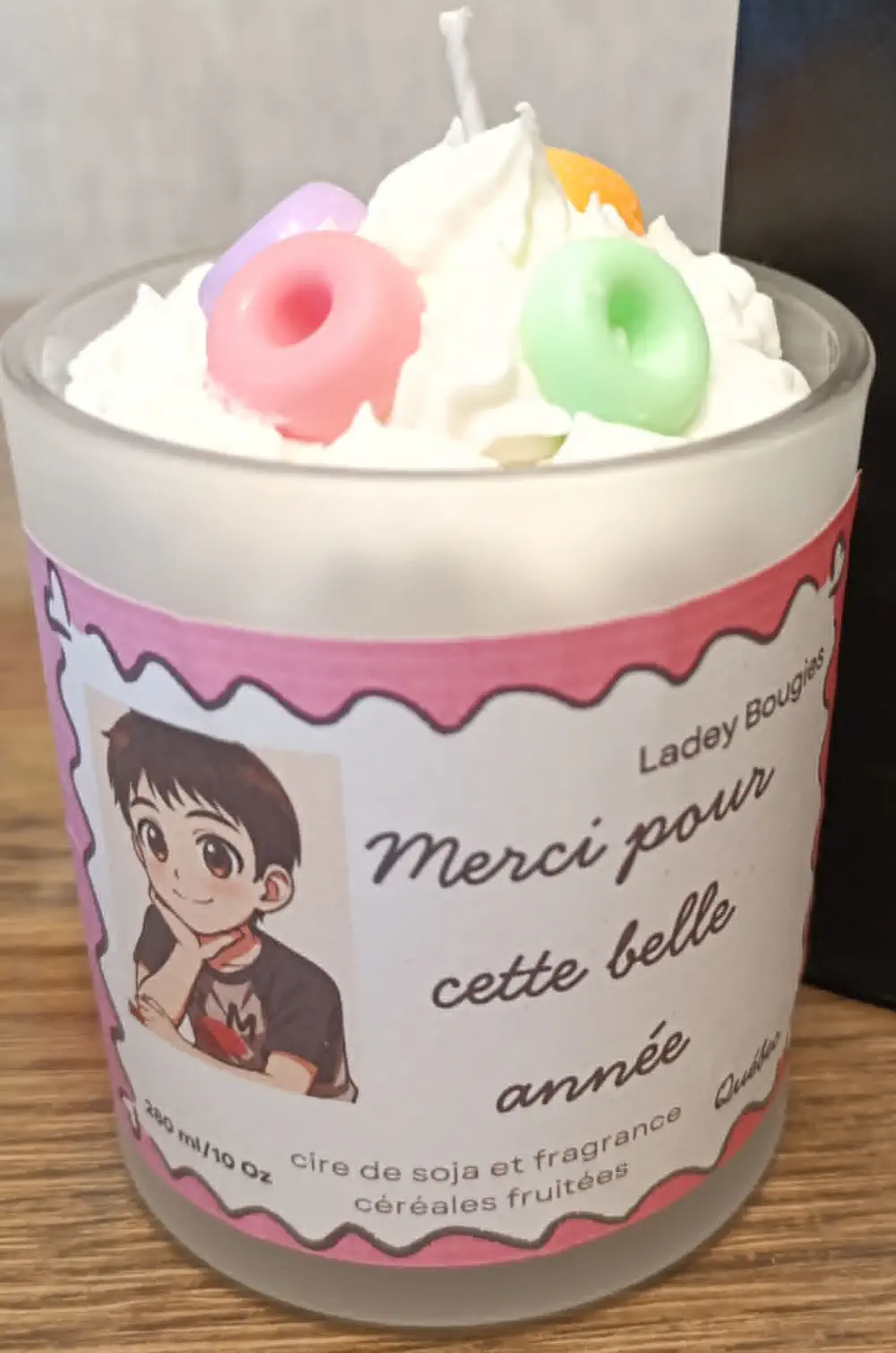 Bougie artisanale dans un verre transparent de 280ml, au parfum céréales fruitées, composée d’une cire blanche coiffée d’un effet chantilly, surmontée d’un décor coloré en forme de petits anneaux façon céréales pastel, accompagnée d’une étiquette illustrée au style enfantin portant un message de remerciement.