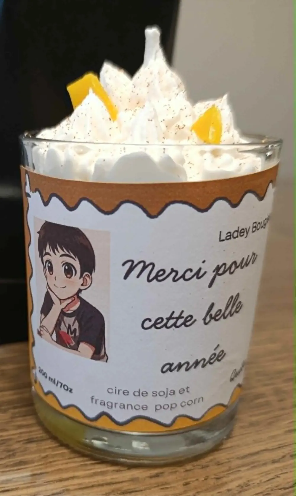 Bougie artisanale dans un verre transparent de 200ml, au parfum pop-corn, composée d’une cire blanche montée en effet chantilly, surmontée de petits éclats jaunes façon friandises, accompagnée d’une étiquette illustrée au style anime portant un message de remerciement.